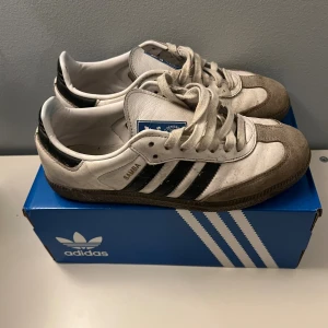 Adidas Samba sneakers - Klassiska Adidas Samba sneakers i vitt skinn med svarta ränder och detaljer i beige mocka. Låg modell med snörning och brun gummisula. Perfekta för dig som gillar att öka din stil till 100%. Ikonisk blå Adidas-logga på plösen.