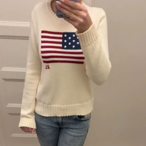 Vit stickad tröja Polo Ralph Lauren - Vit stickad tröja från Polo Ralph Lauren med amerikansk flagga i blått, rött och vitt på bröstet samt RL-broderi. Rund hals och ribbade muddar vid ärmslut och nederkant. Perfekt för dig som gillar klassisk och clean stil.