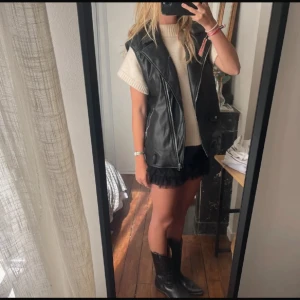 Svart skinnväst med dragkedja - Snygg svart väst i skinnimitation med breda slag och två dragkedjor framtill. Västen har en cool, lite oversized passform och fickor med knappdetalj. Perfekt att slänga över en stickad tröja för en edgy look.