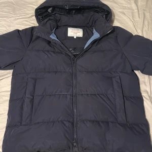 Mörkblå dunjacka från GANT 2XL - Säljer en mörkblå dunjacka från GANT, modell The Alta Down Jacket. Jackan har huva, två stora fickor och dragkedja framtill. Perfekt för kalla dagar med sin puffiga design och stilrena look. GANT-logga på bröstet och patch på ärmen. Storlek 2Xl