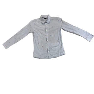 Ljusblå skjorta från Tommy Hilfiger - Snygg ljusblå skjorta från Tommy Hilfiger med små broderade loggor i rött, blått och vitt över hela plagget. Klassisk krage, långärmad och knäppning framtill. Perfekt för dig som gillar stilrena detaljer och ikonisk design.