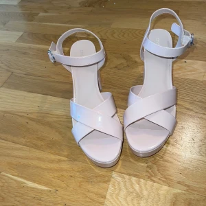 Ljusrosa sandaler med klack från H&M - Säljer ett par ljusrosa sandaler från H&M Divided med bred klack och platåsula. Skorna har korsade remmar framtill, öppen tå och justerbart spänne vid ankeln. Tillverkade i syntetmaterial med en stilren och modern look.