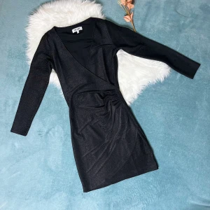 Svart glittrig omlottklänning Dry Lake - Svart kortklänning från Dry Lake med glittrigt tyg och snygg omlott-design. Klänningen har långärmade ärmar, v-ringad hals och draperade detaljer i midjan som ger en cool siluett. Perfekt för dig som vill sticka ut med lite extra glitter.