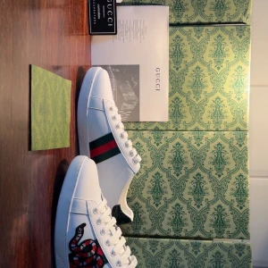 Gucci Ace sneakers med orm  - Gucci Ace sneakers i vitt skinn med klassiska gröna och röda ränder på sidan och broderad orm. Baksidan har metallicdetaljer i rött och grönt med ormskinnsmönster. Snygg och ikonisk modell med platt sula och snörning.