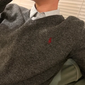 Grå ulltröja från Polo Ralph Lauren - Mörkgrå stickad tröja från Polo Ralph Lauren i 100% lammull. Klassisk v-ringning och röd broderad logga på bröstet. Perfekt att bära över skjorta för en clean och stilren look.