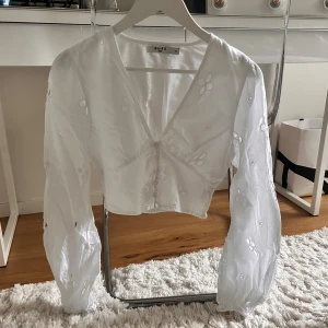 Vit blommig blus från NA-KD - Säljer en söt vit blus från NA-KD i storlek 34. Blusen har broderade blommor, v-ringning och knappar framtill. Den är croppad med långa puffärmar och smock baktill för extra komfort. Perfekt för dig som gillar romantiska detaljer och fräsch stil.