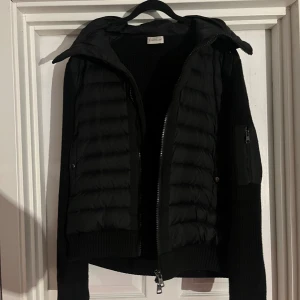 Svart Moncler pufferjacka med huva - Svart pufferjacka från Moncler med stickade ärmar och huva. Jackan har quiltad front, dragkedja och en ficka med dragkedja på ärmen. Snyggt Moncler-märke på armen och ribbade muddar. Perfekt för dig som vill ha en clean och modern look.