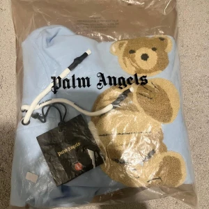 Ljusblå palme Angeles  - Ljusblå hoodie och vit snörning i huvan. Oversized fit med ribbade muddar och känguruficka. Tillverkad i mjuk polyester, perfekt för en chill och trendig look.