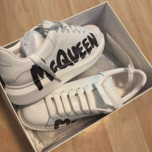 Alexander McQueen vita sneakers - Säljer ett par vita Alexander McQueen sneakers i skinn med chunky sula och svart graffiti-liknande McQueen-tryck på sidan. Klassisk rund tå, bred platt sula och vita skosnören. Kommer i originalkartong med extra skosnören och dustbag.