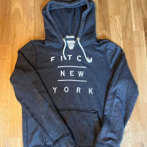 Abercrombie & Fitch hoodie - Mörkgrå hoodie från Abercrombie & Fitch med vit broderad text 'Fitch New York' på bröstet. Tröjan har huva med vita snören och en klassisk känguruficka framtill. Perfekt för chill dagar och snygg streetstil.