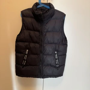 Svart dunväst från Woxo 720° - Svart dunväst från Woxo 720° med hög krage och quiltad design. Västen har två fickor med dragkedja och detaljerade band med logga. Materialet är polyamid och polyester, vilket gör den lätt och bekväm. Perfekt för lager-på-lager under kyliga dagar.