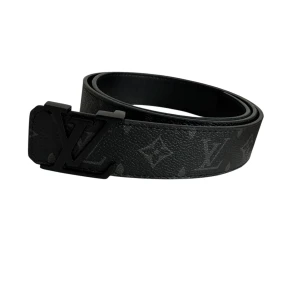 Svart Louis Vuitton bälte med monogram - Snyggt svart bälte från Louis Vuitton med klassiskt monogrammönster och stor LV-logga som spänne. Bältet är gjort i exklusivt skinn och har en mörk, stilren look som passar till jeans eller kostym. Perfekt för dig som gillar lyxiga accessoarer.