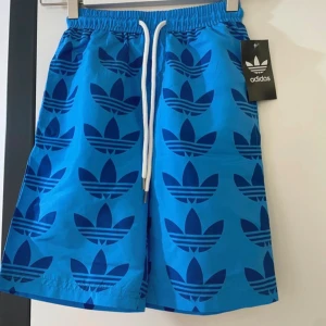 Blå Adidas shorts med snörning - Säljer ett par blå shorts från Adidas med stora mörkblå loggor över hela tyget. De har elastisk midja med vit snörning och är tillverkade i lätt polyester. Perfekta för dig som gillar sportig stil och vill sticka ut med ett fett mönster.shorts är för barn i ålder 4,5