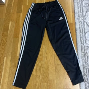 Svarta Adidas träningsbyxor - Svarta träningsbyxor från Adidas med klassiska tre vita ränder längs sidorna och Adidas-logga på låret. Byxorna har elastisk midja med snörning och är gjorda i ett mjukt syntetmaterial som är skönt mot huden. Perfekta för chill eller sport. Fickor i framsidan 