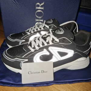 Svarta sneakers från Dior - Snygga svarta sneakers från Dior med vita detaljer och reflekterande logga på sidan. Skorna har vit sula, snörning och meshpartier för extra andningsförmåga. Perfekta för dig som vill sticka ut med exklusiv streetstyle.