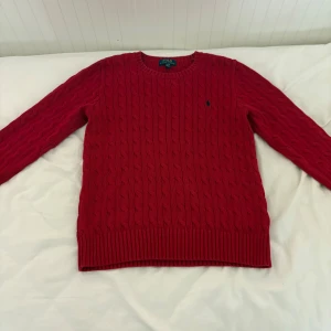 Röd kabelstickad tröja Polo Ralph Lauren - Röd kabelstickad tröja från Polo Ralph Lauren med rund halsringning och diskret broderad logga på bröstet. Tröjan har långa ärmar och ribbade muddar vid ärmslut och nederkant. Perfekt för dig som gillar klassisk och stilren look.