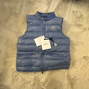 Ljusblå dunväst från Moncler - Snygg ljusblå dunväst från Moncler med hög krage och två dragkedjefickor framtill. Västen har quiltad design, Moncler-logga på bröstet och dubbla dragkedjor. Perfekt för lager-på-lager och ger en sportig vibe. Tags följer med. Skriv för mer info. Passar S.