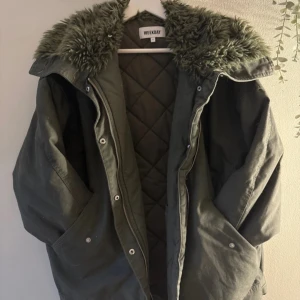 Olivgrön parkas från Weekday med fuskpäls - Säljer en olivgrön parkajacka från Weekday med stor, fluffig fuskpäls på kragen och quiltat foder. Jackan har dragkedja och knappar framtill samt stora fickor. Perfekt för kyliga dagar och riktigt snygg till streetwear-stilen.