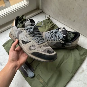 Valentino Rockrunner sneakers grå - Snygga Valentino Rockrunner sneakers i olika grå nyanser med camouflage-mönster och detaljer i mocka och mesh. Skorna har platt sula, rund tå och silverfärgade nitar vid hälen. Medföljer extra skosnören och dustbag.