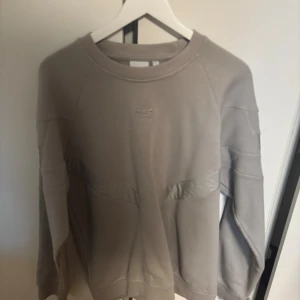  sweatshirt från Adidas - Säljer en beige sweatshirt från Adidas med diskret logga framtill och baktill. Tröjan har långärmad design och rund halsringning, samt snygga sömdetaljer över bröstet och ryggen. Perfekt för dig som gillar stilrena och sportiga plagg.