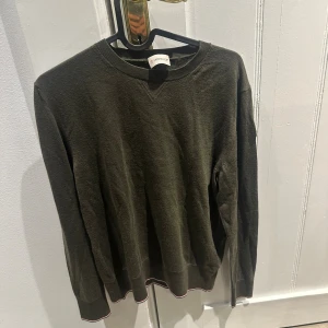 Mörkgrön Moncler stickad tröja - Snygg mörkgrön stickad tröja från Moncler med rund hals och långa ärmar. Tröjan har Moncler-logga på vänster ärm och en diskret röd-vit-blå rand vid nederkanten. Perfekt för dig som gillar stilrena och exklusiva plagg.