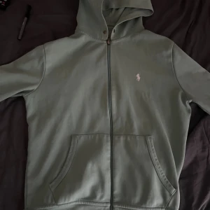 Grön hoodie från Polo Ralph Lauren - Snygg grön hoodie från Polo Ralph Lauren med klassisk vit logga på bröstet. Tröjan har huva med dragsko, hel dragkedja framtill och två stora fickor. Tillverkad i mjuk bomull för skön känsla. Perfekt för chill dagar och streetwear-stil.