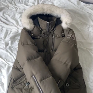Moose Knuckles mörkgrön pufferjacka - Säljer en fräsch äkta mörkgrön pufferjacka från Moose Knuckles med fluffig vit päls på huvan, jackan är i storlek M, snygga metallpatches på ärmen och flera dragkedjor. Jackan har hög krage, långa ärmar och stora fickor med både knapp och zip. Perfekt för kalla vinterdagar. Finns i Stockholm kan även fraktas