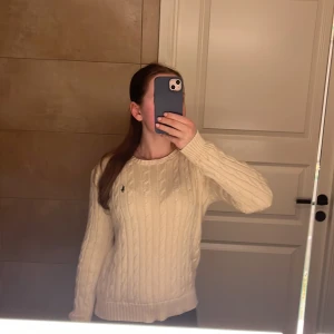 Beige stickad tröja från Polo Ralph Lauren - Säljer en beige stickad tröja från Polo Ralph Lauren i storlek M. Tröjan har klassiskt kabelstickat mönster, rund hals och ribbade muddar. Den har en liten broderad logga på bröstet och är långärmad. Perfekt för dig som gillar stilrena och tidlösa plagg. Knappt använd