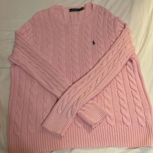 Rosa stickad tröja från Polo Ralph Lauren - Säljer en ljusrosa stickad tröja från Polo Ralph Lauren i storlek L. Tröjan har klassiskt kabelstickat mönster, rund hals och långärmade ribbade muddar. Den ikoniska blå Polo-loggan är broderad på bröstet. Perfekt för dig som gillar preppy stil.