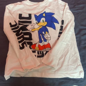 Vit långärmad Sonic-tröja - Cool långärmad tröja i vitt med stort tryck av Sonic the Hedgehog i blått, rött och gult på framsidan. Svart text längs ena ärmen ger extra streetstyle-vibe. Perfekt för dig som älskar gaming och vill sticka ut med en unik look.
