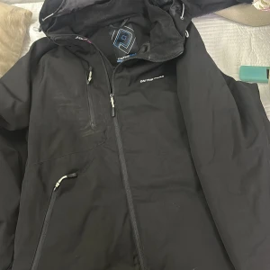 Svart vindjacka från On The Peak - Svart vindjacka från On The Peak med huva och dragkedja framtill. Jackan har flera fickor med dragkedja och snygga detaljer som snörning vid huvan. Insidan är fodrad med mönstrat tyg och jackan har en sportig look som passar perfekt för utomhusaktiviteter.