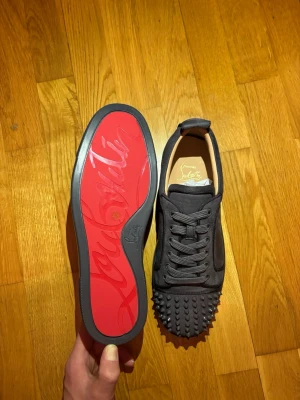 Grå Loubitin sneakers med nitar - Säljer ett par grå sneakers från Christian Louboutin med ikonisk röd sula och coola nitar på tån. Skorna har snörning och är gjorda i mocka med läderdetaljer. Medföljer dustbag, extra nitar och äkthetsbevis. Perfekt för dig som vill sticka ut med lyxig streetstyle. 
