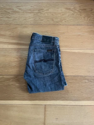 Grå jeans från Nudie Jeans, W30 L34 - Säljer ett par grå jeans från Nudie Jeans i storlek W30 L34. Jeansen har klassisk femficksdesign, svarta detaljer och en snygg tvättad look. Materialet är jeans och passformen är rak med normal midja. Perfekt för dig som gillar stilrena och bekväma jeans. Byxorna är använda få gånger och saknar defekter 