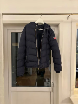 Mörkblå pufferjacka från Vinson - Mörkblå pufferjacka från Vinson med huva och guldfärgad dragkedja. Jackan har ett broderat Vinson-märke på bröstet och en amerikansk flagga på ärmen. Perfekt för kalla dagar och har en schysst, sportig look.