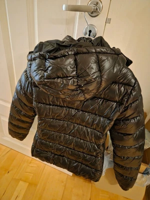 Svart pufferjacka med huva - Säljer en svart glansig pufferjacka med avtagbar huva. Dragkedja på båda fickorna. Jackan är aldrig använd! (råkade köpa två styckna)