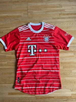 Röd Bayern München adidas tröja - Säljer en röd Bayern München matchtröja från adidas med vita ränder och logga på bröstet. Tröjan har rund hals, korta ärmar och vita detaljer vid krage och ärmslut. Perfekt för dig som älskar fotboll och vill visa ditt stöd för laget.