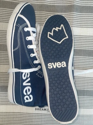 Blå sneakers från Svea, strl 40 - Snygga blå sneakers från Svea med vita detaljer och snörning. Skorna har en vit sula och coolt tryck med Svea-logga på sidan och sulan. Insidan är fodrad med rosa och marinblåa ränder för en extra pop av färg. Perfekta för dig som gillar en sportig och stilren look.