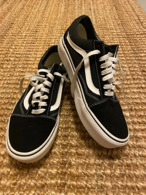 Svarta Vans Old Skool sneakers - Säljer ett par klassiska Vans Old Skool sneakers i svart och vitt med den ikoniska sidostripen. Skorna har vit snörning, gummisula med rutmönster och ovandel i canvas och mocka. Perfekt för dig som gillar streetstyle och skatekänsla. Tjock sula platå.