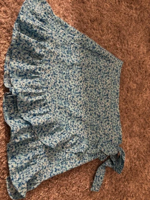 Blommig blå kjol från New Yorker, storlek M - Säljer en kort kjol från New Yorker i storlek M. Kjolen är blå med blommigt mönster och har en volangkant nedtill samt knytband i midjan. Perfekt för dig som gillar färgglada och somriga plagg med lite extra detaljer.