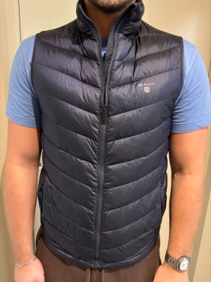 Mörkblå dunväst från GANT - Snygg mörkblå dunväst från GANT, modell Airlight Vest. Västen har quiltad design, hög krage och dragkedja framtill. Perfekt att slänga över en hoodie eller t-shirt för extra värme. Diskret GANT-logga med emblem på bröstet.