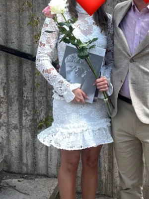 Vit spetsklänning med volang studentklänning - Superfin vit kort spetsklänning med blommigt mönster och volang nertill. Klänningen har långärmade ärmar och hög hals, samt fina detaljer i spets och volang. Perfekt för student eller sommarens tillfällen!