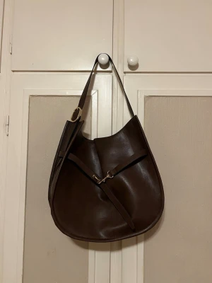 Brun axelväska  - Säljer denna jättefina axelväska från H&M då jag har köpt en annan väska! I nyskick då den bara är använd 1-2 gånger med en rymlig insida med ett litet innerfack, perfekt att ha som skolväska!