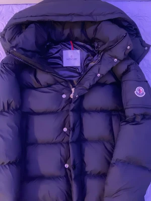 Svart Moncler Vezere - Säljer en svart dunjacka från Moncler med stor, mysig huva och klassisk logga på ärmen. Jackan har quiltad design, dragkedja och knappar framtill. Materialet är glansigt och känns riktigt lyxigt, perfekt för kalla dagar. Passformen är normal och jackan är långärmad.