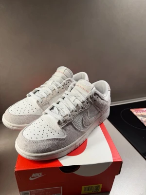 Nike Dunk Low Summit White Photon Dust Univeristy Red - Nike Dunk Low sneakers i färgen Summit White/Photon Dust. Skorna har en snygg mix av slät och ruggad mocka samt läder, med perforerad tå och klassisk snörning. Låg modell med vit och ljusgrå ton, diskret röd detalj på plösen och broderad Nike-logga på hälen.                          Helt nya, säljes pga för liten storlek för mig