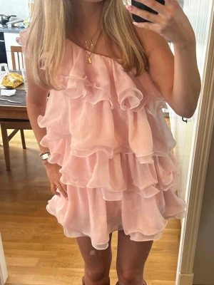 Rosa volangklänning med oneshoulder - Supersöt rosa kortklänning med flera lager volanger och oneshoulder-design. Klänningen är ärmlös och har en luftig, flowy känsla tack vare det tunna materialet. Perfekt för dig som gillar romantisk och lekfull stil.