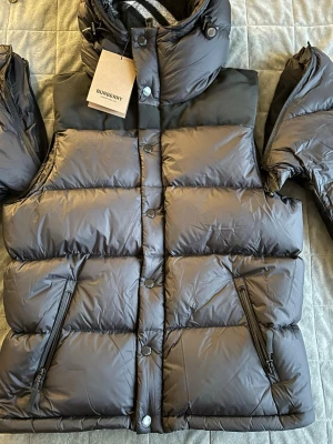 Svart pufferjacka från Burberry - Säljer en svart pufferjacka från Burberry med avtagbara ärmar så den kan användas som väst. Jackan har huva med klassiskt rutigt foder, dragkedja och knappar framtill samt två sidofickor med dragkedja. Jackan är i storlek L och passar Perfekt för kalla dagar och riktigt snygg stil.