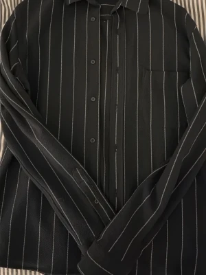 Navy randig skjorta ZARA - Snygg svart skjorta med vita vertikala ränder och klassisk krage. Skjortan har långa ärmar, knäppning framtill och en bröstficka. Materialet känns lite strukturerat och ger en cool vibe. Perfekt för dig som gillar stilrena och moderna plagg. Från Zara.