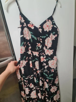 Blommig byxdress från Amisu, strl 36 - Säljer en svart byxdress från Amisu i storlek 36 med blommigt mönster i ljusa toner och gröna blad. Dressen har smala axelband, omlott upptill och ett medföljande bälte i samma tyg. Materialet är mjukt och lätt, perfekt för varma dagar.