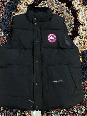 Svart dunväst från Canada Goose - Säljer en svart dunväst från Canada Goose med hög krage och tryckknappar framtill. Västen har två stora fickor med logotyp på ena fickan och ett klassiskt rundat märke på bröstet. Perfekt för kyliga dagar och enkel att matcha med olika outfits.