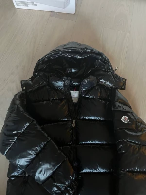 Svart Moncler pufferjacka med huva - Säljer en svart pufferjacka från Moncler med glansig finish och huva. Jackan har Moncler-logga på ärmen, dragkedja framtill och quiltad design. Perfekt för kalla dagar och har en riktigt snygg streetstil. Materialet är polyester och dun.